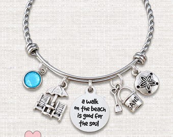 May include: A silver-toned twisted wire charm bracelet featuring a round blue crystal, a beach chair and umbrella charm, a circular pendant engraved with the text a walk on the beach is good for the soul, a shovel and sand bucket, and a sand dollar.