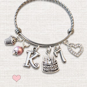 May include: A silver-toned twisted wire bangle bracelet featuring multiple charms: a cupcake, a pink pearl bead, the letter K, a birthday cake charm with the text HAPPY BIRTHDAY, the number 7, and a heart-shaped charm encrusted with clear crystals.