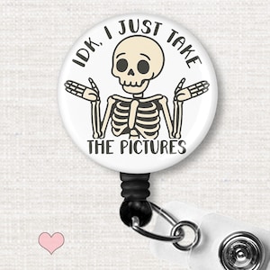 May include: A circular badge reel featuring a cartoon skeleton shrugging with the text IDK, I JUST TAKE THE PICTURES in black lettering. The skeleton is beige with dark outlines, set against a white background on a black retractable cord.