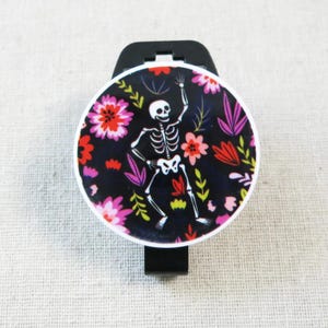 May include: A black phone grip with a round, floral design featuring a dancing skeleton. The design includes pink, red, and purple flowers on a black background. The grip has a white border.