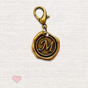 May include: A bronze-toned metal charm featuring a circular wax seal design with an embossed letter M in the center. The charm is attached to a lobster claw clasp, creating a functional accessory piece with a metallic finish.