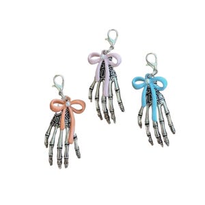 May include: Three silver-toned skeleton hand charms, each featuring a small white bead and a bow in either peach, light pink, or light blue. Each charm includes a silver metal lobster clasp at the top for attachment to various items.