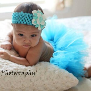 May include: A baby girl wearing a light blue tulle tutu and a headband with a white flower. The baby is lying on a white fluffy surface and looking at the camera.