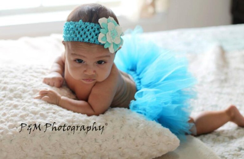 May include: A baby girl wearing a light blue tulle tutu and a headband with a white flower. The baby is lying on a white fluffy surface and looking at the camera.