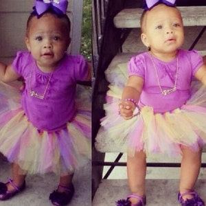 May include: A young girl wearing a purple shirt and a multi-colored tulle skirt. She is wearing a purple bow in her hair and a silver necklace. She is standing on a set of stairs.