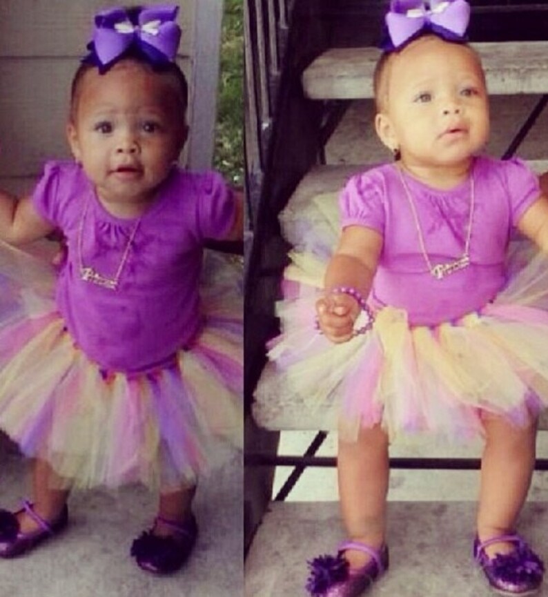 May include: A young girl wearing a purple shirt and a multi-colored tulle skirt. She is wearing a purple bow in her hair and a silver necklace. She is standing on a set of stairs.
