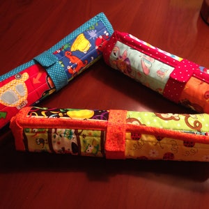 Peut inclure: Trois étuis à crayons en tissu colorés avec des motifs différents. Les étuis sont enroulés et présentent une palette de couleurs rouge, orange, jaune, vert, bleu et blanc. Les étuis sont en tissu matelassé et présentent une variété de motifs d'animaux et de véhicules.