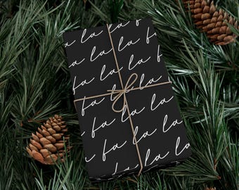 May include: A rectangular gift box wrapped in black paper featuring a repeating white cursive script pattern of the words fa la la. The box is tied with thin brown twine arranged in a cross pattern and secured with a simple bow at the center.