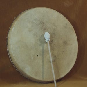 May include: A large, round drum with a light tan drumhead and a white drumstick. The drumhead has a slightly textured appearance. The drumstick has a rounded head. The drum is set against a brown background.