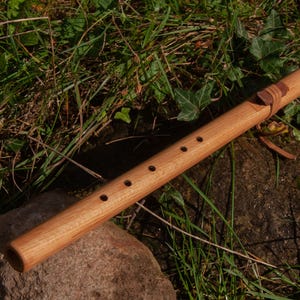 May include: A handcrafted wooden flute with a natural finish, featuring six finger holes and a leather accent. The instrument rests on a stone amidst green foliage, showcasing its rustic charm. This flute is approximately 46 cm long.
