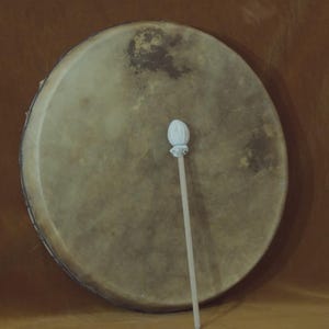 May include: A large, round drum with a natural hide surface and a white drumstick. The drum has a light brown color with darker spots and is set against a brown background. The drumstick has a white, rounded head.