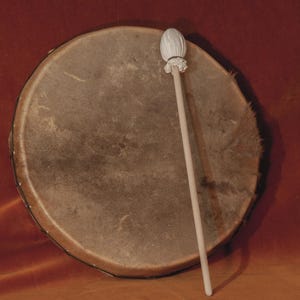 May include: A round, brown drum with a light-colored drumstick resting on its surface. The drum has a natural, textured appearance, and the drumstick has a rounded head. The background is a solid, reddish-brown color.