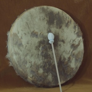May include: A large, round drum with a natural hide surface, featuring mottled brown and gray tones. A white-tipped drumstick rests diagonally across the drum's face, suggesting it's ready for use. The drum's rim is dark, and the background is a warm brown.