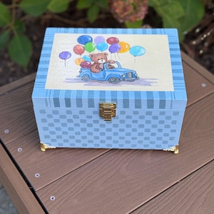 May include: A light blue wooden box with a checkered pattern and gold-colored feet. The lid features a watercolor painting of a teddy bear driving a blue truck filled with colorful balloons. The box has a gold-colored clasp.