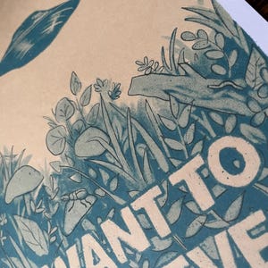 May include: Illustration of a forest scene with the words "I WANT TO BELIEVE" in white block letters. The artwork features stylized plants, insects, and a leaf in shades of blue and green on a beige background.