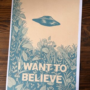 May include: A print featuring a blue UFO hovering above a detailed illustration of plants and foliage. The words "I WANT TO BELIEVE" are printed in large white letters at the bottom of the image. The background is a light tan color.