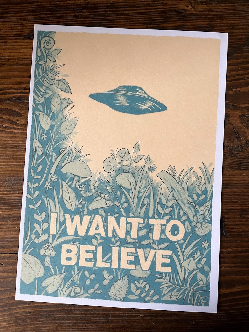 May include: A print featuring a blue UFO hovering above a detailed illustration of plants and foliage. The words "I WANT TO BELIEVE" are printed in large white letters at the bottom of the image. The background is a light tan color.