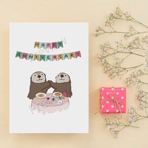 May include: A white card with the text "Happy Anniversary" in colorful letters with a banner above it. Two otters are sitting at a table with a plate of food, a glass of wine, and a rose.
