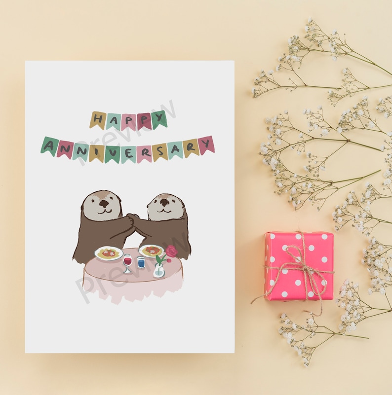 May include: A white card with the text "Happy Anniversary" in colorful letters with a banner above it. Two otters are sitting at a table with a plate of food, a glass of wine, and a rose.