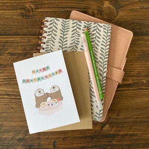 May include: A card with a happy anniversary message and a cute illustration of two otters holding hands at a table with food and drinks. The card is on top of a brown envelope and a pink and gray notebook with a green pen.