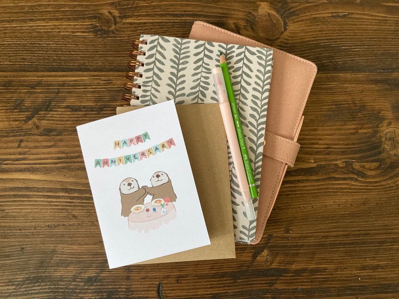 May include: A card with a happy anniversary message and a cute illustration of two otters holding hands at a table with food and drinks. The card is on top of a brown envelope and a pink and gray notebook with a green pen.