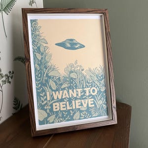 May include: Framed art print featuring a blue UFO above a field of stylized plants and the text "I WANT TO BELIEVE" in white. The print has a cream-colored background and is set in a dark brown wooden frame. The artwork is displayed on a wooden surface.