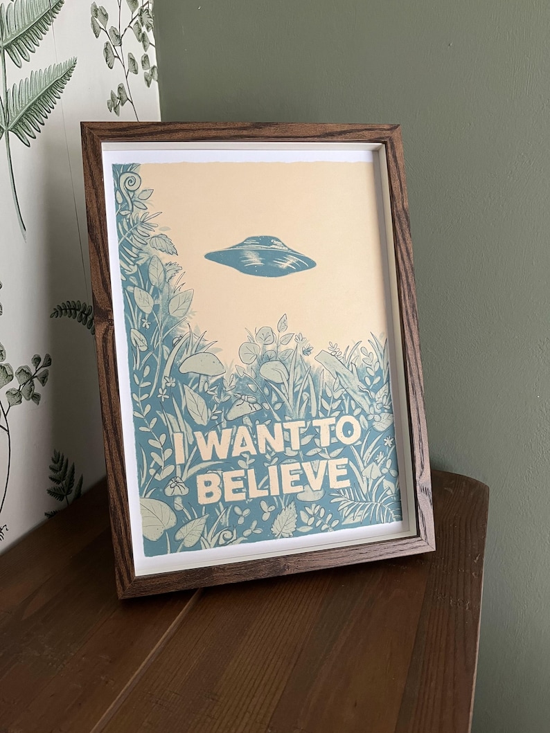 May include: Framed art print featuring a blue UFO above a field of stylized plants and the text "I WANT TO BELIEVE" in white. The print has a cream-colored background and is set in a dark brown wooden frame. The artwork is displayed on a wooden surface.