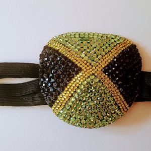 May include: An eye patch featuring a design inspired by the Jamaican flag, crafted with black, yellow, and green rhinestones arranged in a diagonal cross pattern. The patch is attached to a black elastic strap with an adjustable sliding buckle.