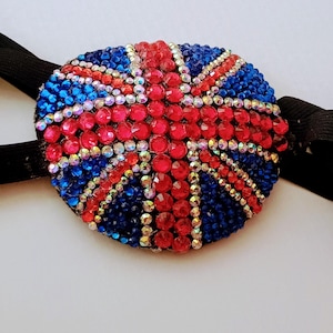 May include: A circular eye patch featuring a Union Jack flag design created with red, blue, and clear rhinestones. The patch is attached to a black elastic strap with adjustable metal hardware, designed to secure the piece comfortably in place.