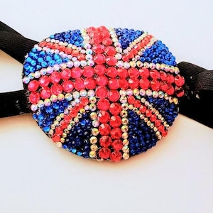 May include: A circular eye patch featuring a Union Jack flag design created with red, blue, and clear faceted rhinestones. The patch is attached to a black elastic strap with a metal adjustment buckle, designed to secure the piece in place.