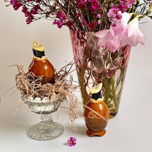 May include: A still life featuring a pink-tinted glass vase filled with a vibrant bouquet of flowers in shades of pink, green, and orange. Two decorative ceramic bird figurines are displayed, one in a glass pedestal bowl with a nest, and the other standing alone.