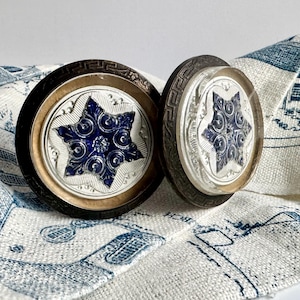 May include: Two circular drawer knobs featuring a dark brown outer rim with a Greek key pattern, a silver-toned middle ring, and a central clear glass insert displaying a raised, deep blue star-shaped emblem with intricate swirling embossed details.