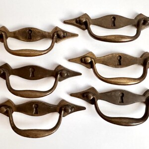 May include: A set of six antique brass drawer pulls, each featuring a horizontal backplate with pointed ends and a central keyhole cutout. A curved, swinging handle is attached to each backplate, providing a functional grip for furniture.