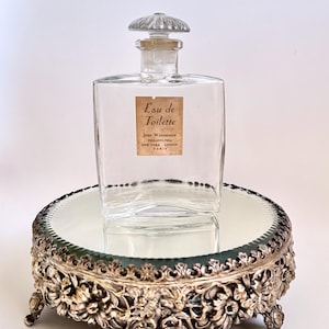 May include: A clear glass perfume bottle with a decorative stopper, labeled "Eau de Toilette." The bottle sits on a mirrored, ornate silver-toned pedestal with floral detailing. The pedestal has three small feet.