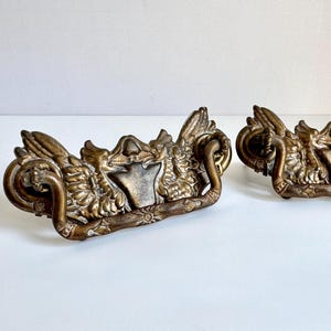 May include: Two matching brass drawer pulls featuring an ornate, winged design with a central shield-like emblem and a curved, textured drop handle. The metal has a darkened, aged patina with intricate relief detailing across the entire surface.