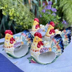May include: A set of six ceramic napkin rings shaped like colorful chickens. Each chicken has a red comb, yellow body, and blue and yellow wings. The napkin rings are on a floral tablecloth.