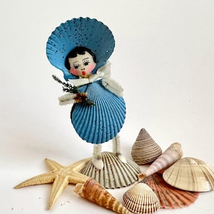 May include: A whimsical seashell doll with a blue shell dress and hat, holding a sprig of greenery. Various seashells and a starfish are arranged around the doll, creating a coastal theme. The doll's face is hand-painted with delicate features.