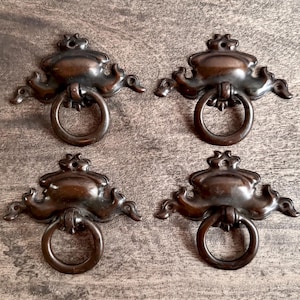 May include: Four antique bronze drawer pulls with ring handles. Each pull has an ornate, decorative design with curved edges and a circular ring handle. The pulls are arranged on a wooden surface.
