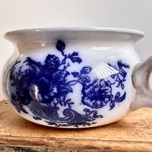 May include: A white and blue ceramic bowl with a handle. The bowl features a blue floral pattern and a wide rim. The handle is curved and attached to the side of the bowl. The bowl is sitting on a wooden surface.
