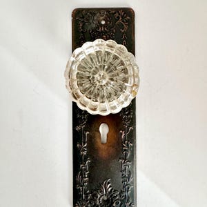 May include: A clear, fluted glass door knob attached to a rectangular, dark bronze-toned metal backplate. The backplate features an intricate, embossed floral and vine pattern with a central keyhole opening and screw holes at the top and bottom.