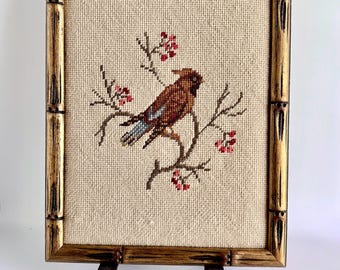 May include: Cross-stitch artwork of a bird perched on a branch with red berries. The artwork is framed in a bamboo-style gold frame and displayed on a metal stand. The bird has brown and blue feathers.
