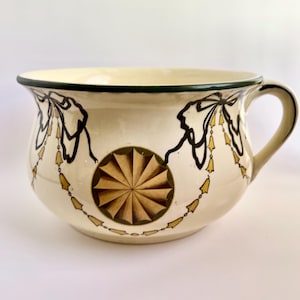 May include: Cream-colored ceramic bowl with a handle, featuring a decorative design. The bowl has a green rim and is adorned with black and gold accents, including a central starburst pattern and garland-like motifs.