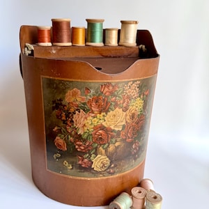 May include: A vintage sewing box with a floral print and a collection of wooden spools of thread. The box is cylindrical and brown, with a curved top. The spools are various colors, including orange, brown, green, and cream.