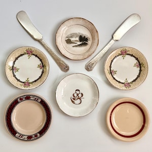 May include: An overhead shot of a collection of vintage small plates and butter knives. The plates feature floral, landscape, and geometric designs in various colors. Two silver butter knives are positioned on either side of the plates.