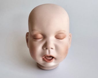 May include: A pale, smooth-textured doll head with a bald scalp, closed eyes, and a slightly open mouth revealing a small tongue. The facial features are painted with soft pinkish tones on the cheeks and lips, creating a realistic appearance.