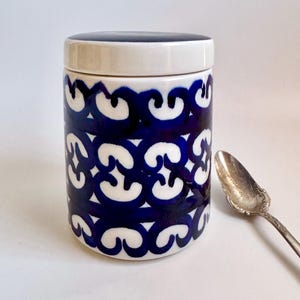 May include: A ceramic jar with a lid, featuring a repeating pattern of blue and white swirls. The jar is cylindrical and sits next to a silver spoon. The lid is dark blue.