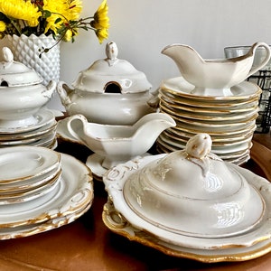 May include: A white porcelain dinnerware set with gold trim. The set includes a covered serving dish, a gravy boat, a covered casserole dish, and several plates.