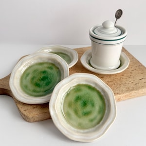 May include: A set of three small, round, ceramic dishes with a wavy edge and a glossy green crackle glaze finish, accompanied by a white ceramic lidded jar with a dark green rim and a small metal spoon resting inside the lid.