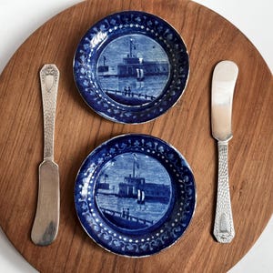 May include: Two blue and white decorative plates with a harbor scene, and two silver butter knives are arranged on a wooden serving board. The plates have a detailed border design. The knives have ornate handles.
