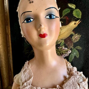 May include: A vintage mannequin head and shoulders featuring a smooth, pale complexion, painted blue eyes, arched black eyebrows, and bright red lips. The mannequin wears a beige lace garment around the neck and shoulders, with a small metal pin visible on the forehead.